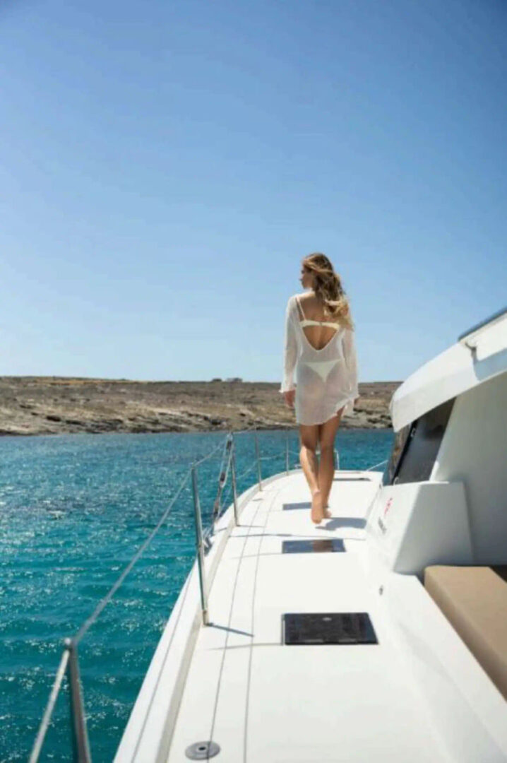 Woman walking along the deck of the Fountaine Pajot 47 catamaran with clear blue waters of Mykonos in the background, a cruise by Events on Board.